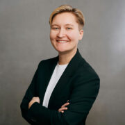 Salome posing with their arms crossed in front of a neutral background. They're wearing a black blazer and have short blond hair.